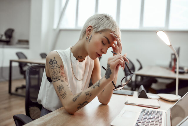 a woman with tattoos on her arm is stressed while sitting in front of a laptop and thinking about how to rebalance employee well being.