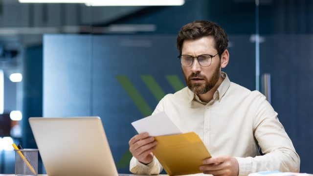 Upset young businessman man sitting in the office at the table and reading the received letter, disappointed with the news and results