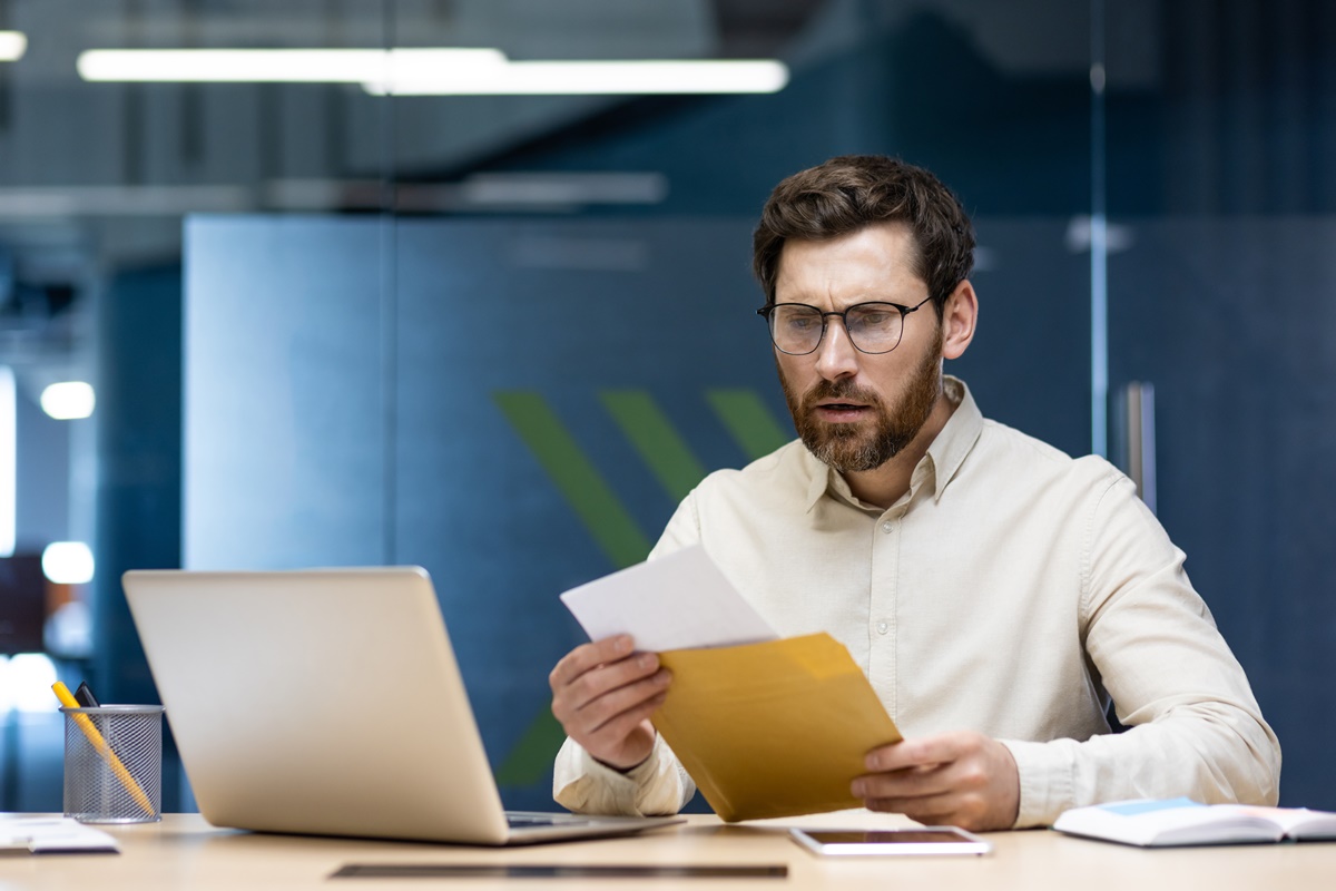 Upset young businessman man sitting in the office at the table and reading the received letter, disappointed with the news and results