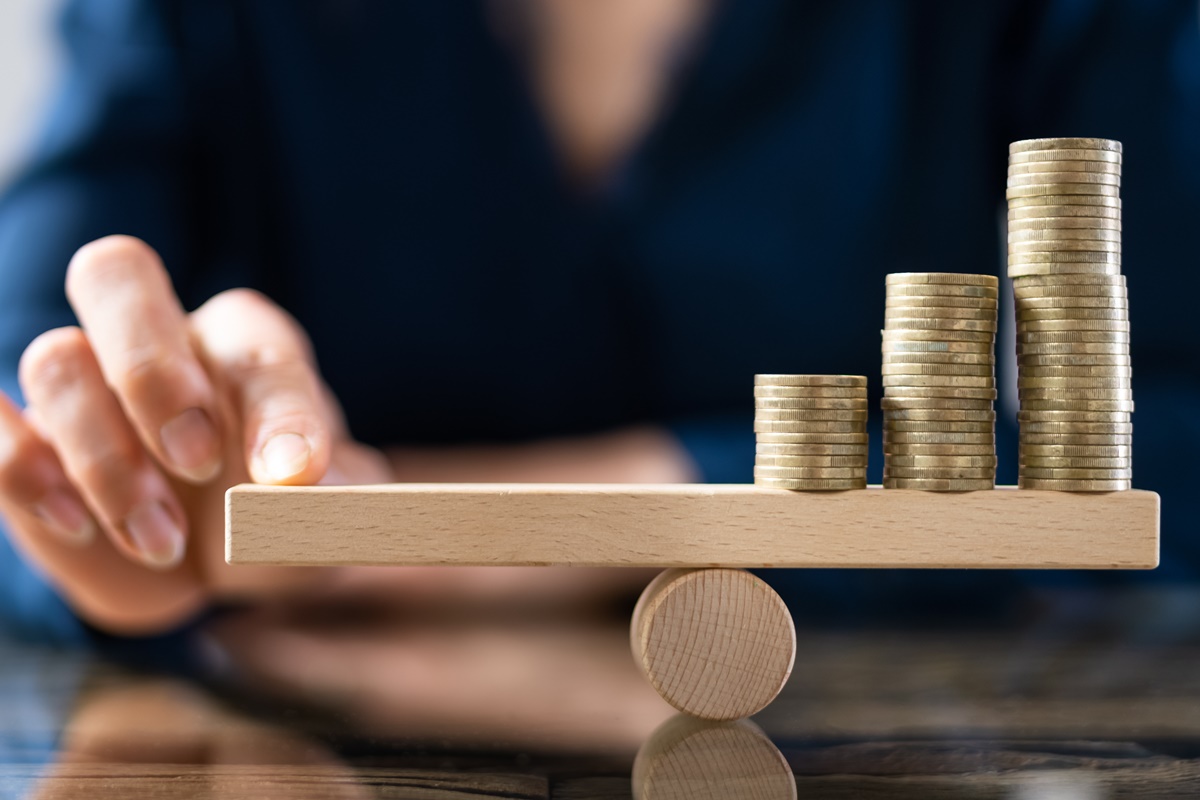 Close-up Of A Businessperson Balancing Stacked Coins On Seesaw With Finger