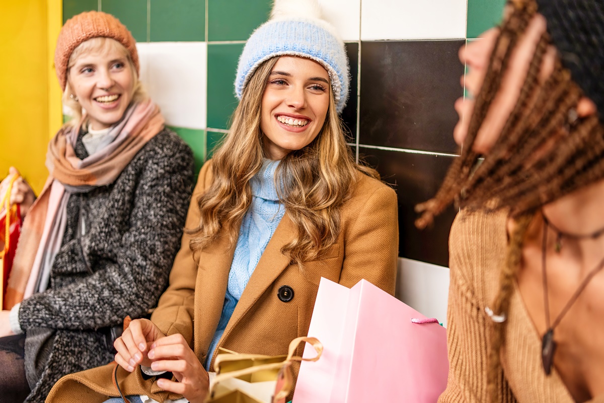 Group of diverse young women friends at subway station after winter shopping. Cheerful millennials in colorful warm clothes, holding bags, socializing while waiting