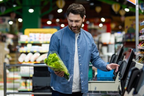 Man using self-checkout machine in grocery store, holding fresh lettuce. Depicts modern shopping experience, convenience, and efficiency. Reflects contemporary lifestyle choices supermarket setting.