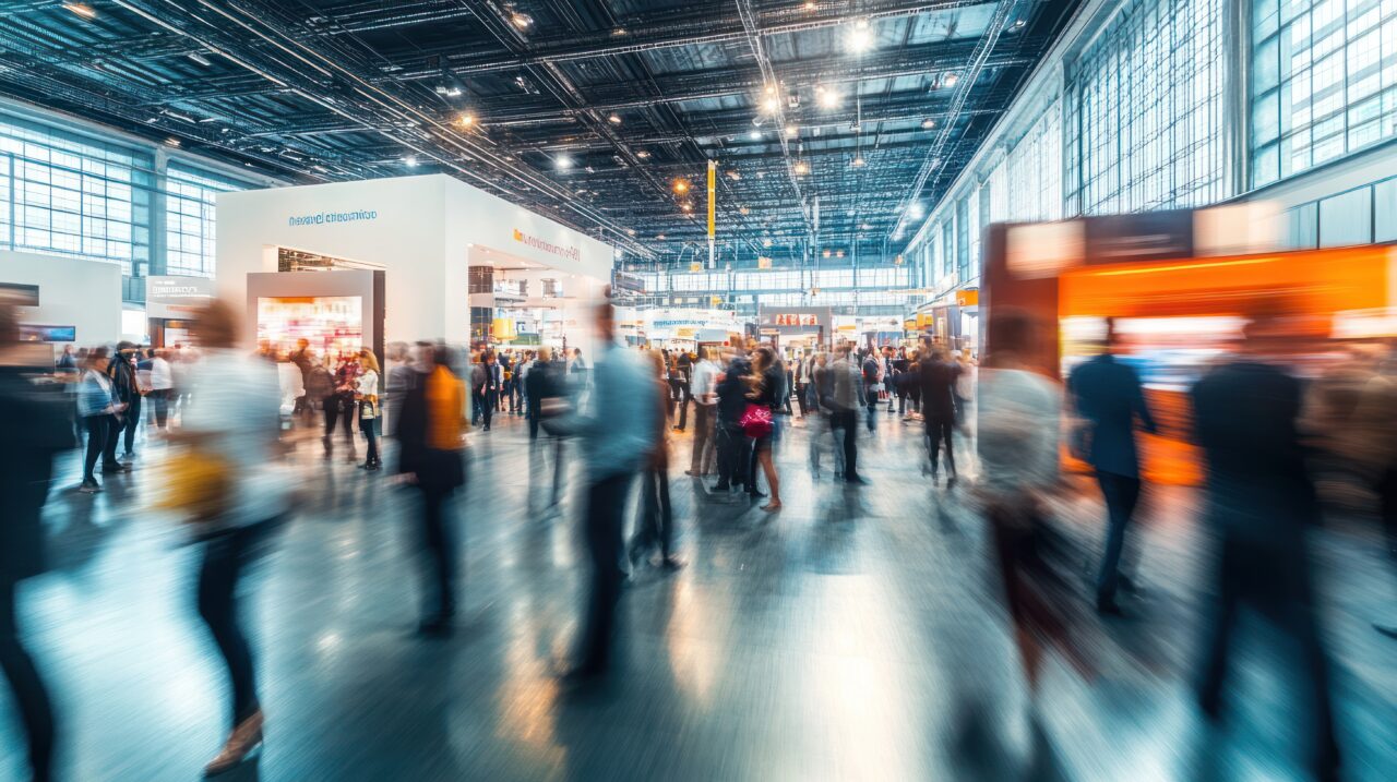 A dynamic scene at a busy trade fair with blurred figures of people networking and interacting around modern exhibition booths, capturing the energy and movement of a vibrant business event