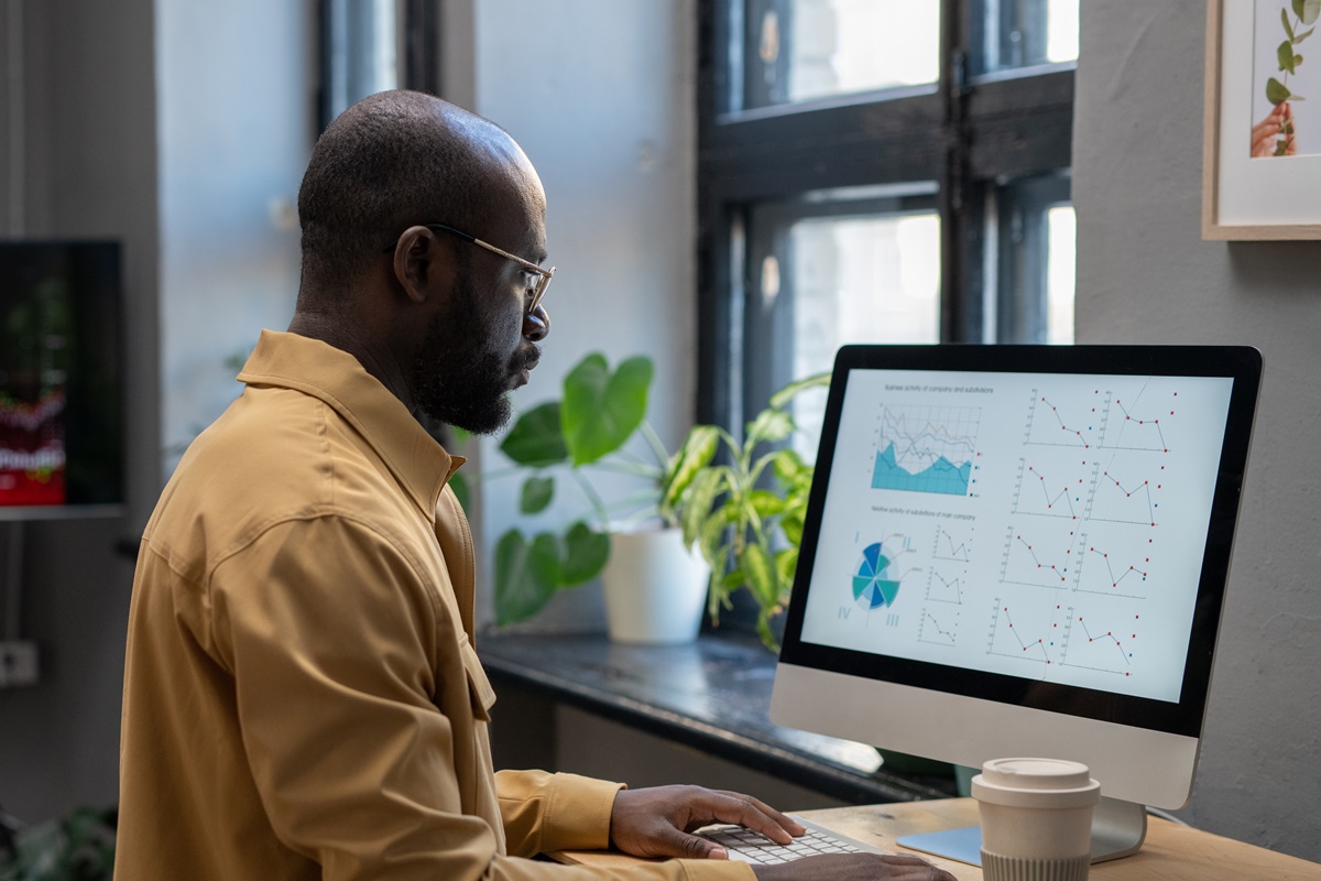 Young modern economist in casual clothes working with financial data in front of computer while sitting by desk in office