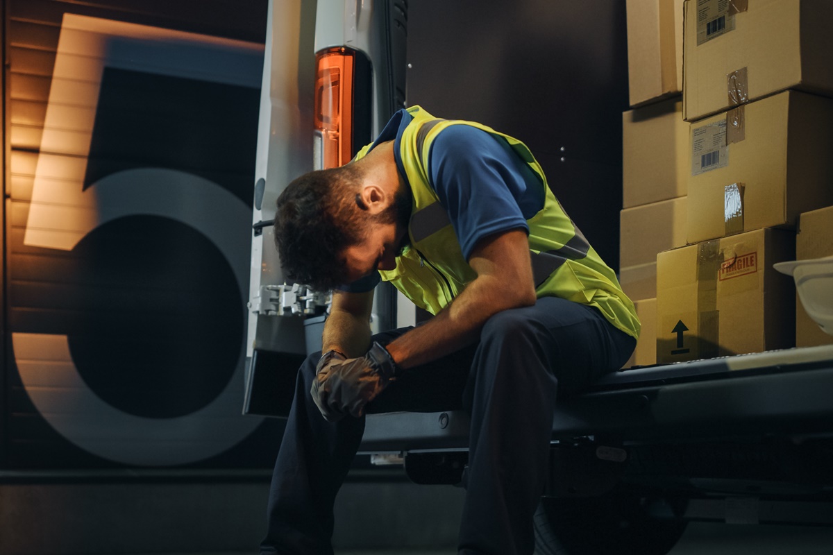 Male Worker Wearing Hard Hat Loads Cardboard Boxes into Delivery Truck, Rests. Online Orders, E-Commerce Goods, Food, Medicine. Tired Overworked Frontline Hero. Dramatic Shot