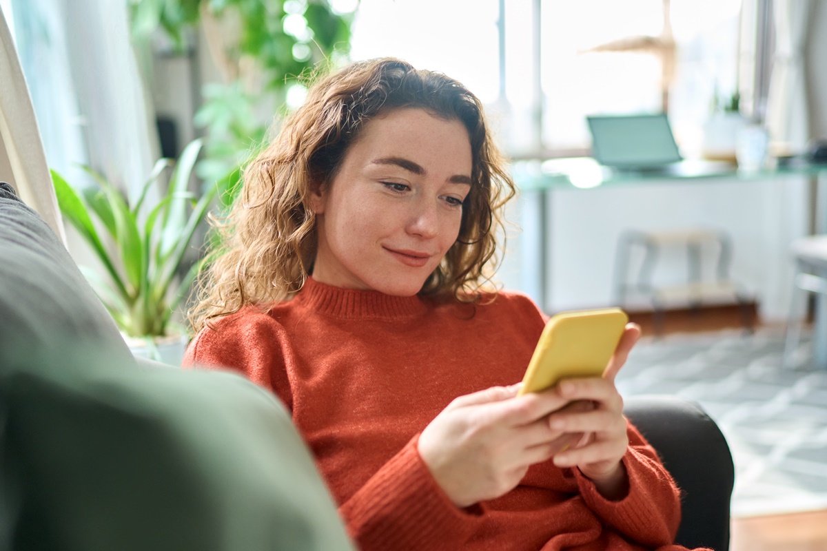 Smiling relaxed young woman sitting on couch using cell phone technology, happy lady holding smartphone, scrolling, looking at cellphone enjoying doing online ecommerce shopping in mobile apps.