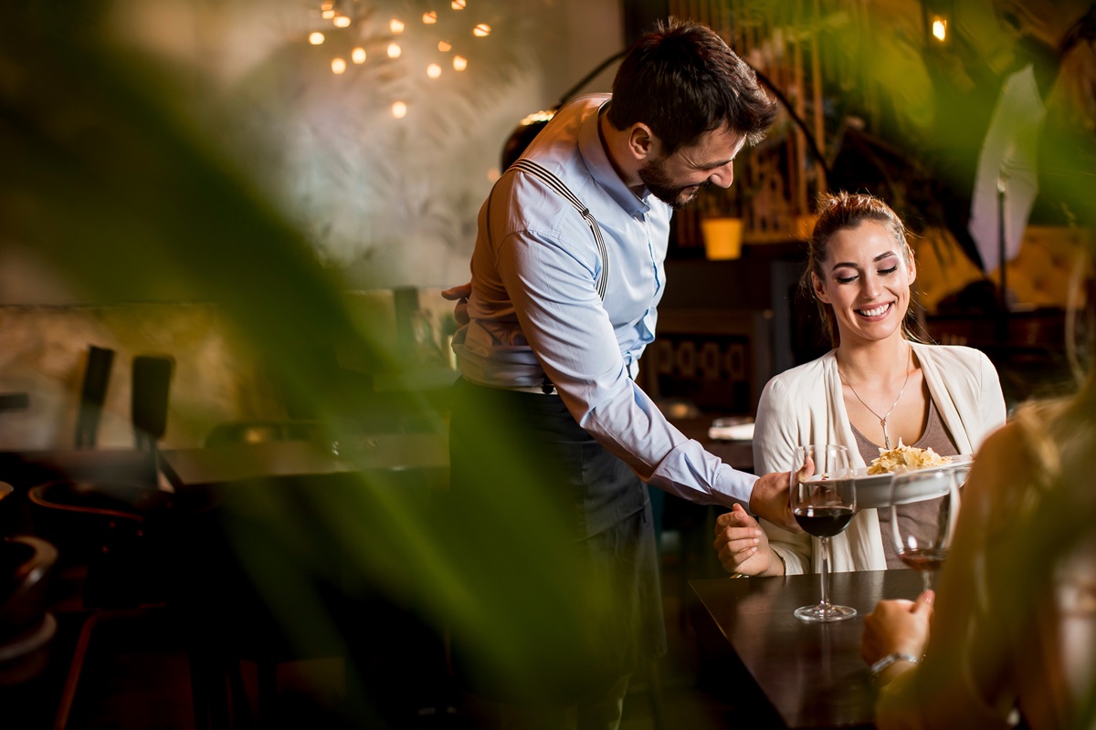 Two smiling young female friends at a restaurant with waiter serving dinner