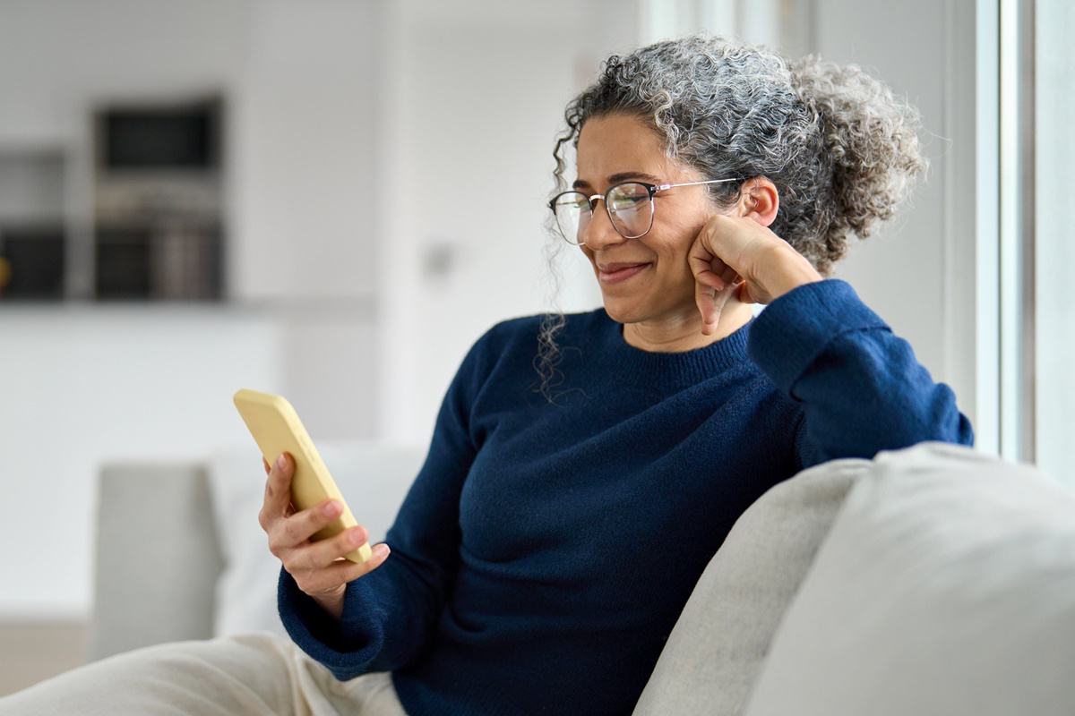 Happy middle aged older woman using smartphone sitting on couch at home. Smiling mature lady holding cellphone browsing internet, texting messages, doing online shopping on mobile cell phone on sofa.