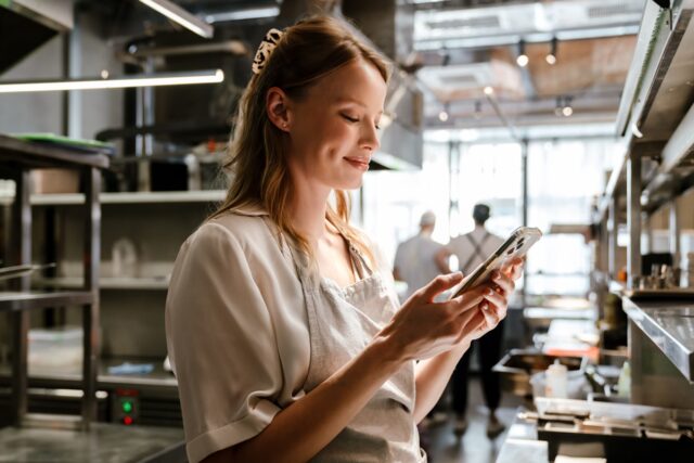 Smiling young woman cook wearing apron using mobile phone while standing in kitchen of a restaurant