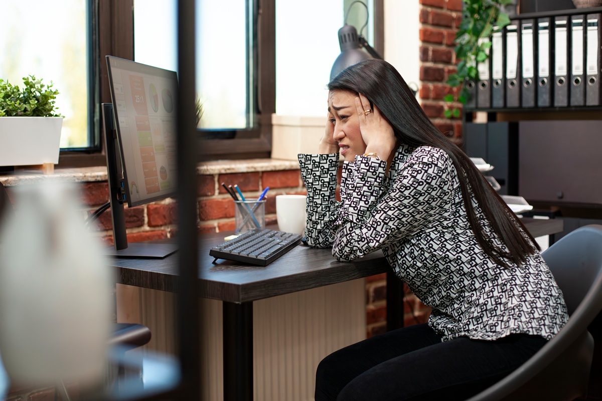 Frustrated female manager leaning on table, overwhelmed by marketing strategy preparation on computer. Asian businesswoman holds forehead in hands at desk, experiencing a headache in modern workspace.