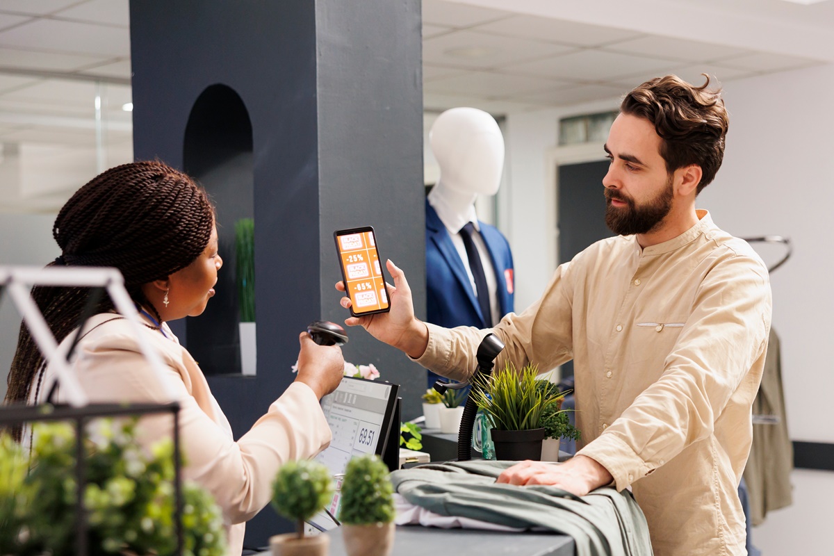 African American woman cashier scanning Black Friday mobile promo code from customer smartphone, using barcode scanner. Young man shopper using coupon to get discount while shopping in clothing store